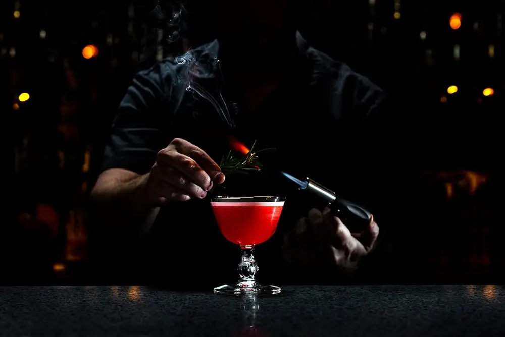 A bartender using a torch to smoke rosemary over a red cocktail, creating a dramatic and smoky effect.