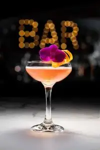 A floral pink cocktail garnished with a vibrant flower and orange twist, set against a bokeh background spelling "BAR".