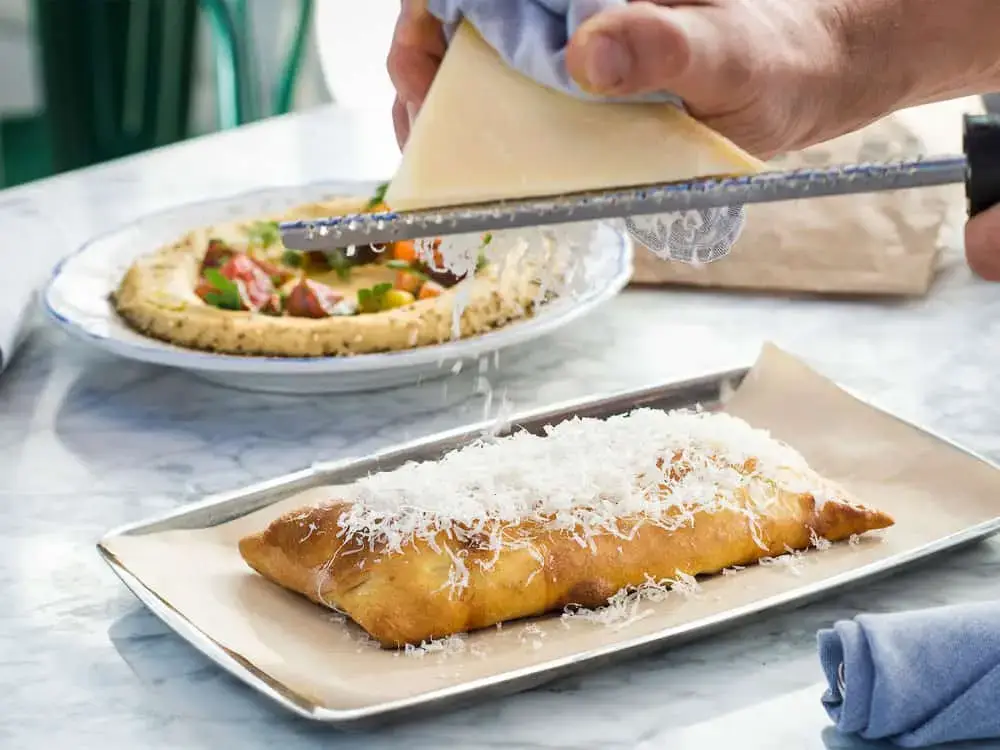 Freshly grated cheese being added to a hot stromboli on a metal tray, with a plate of hummus in the background.