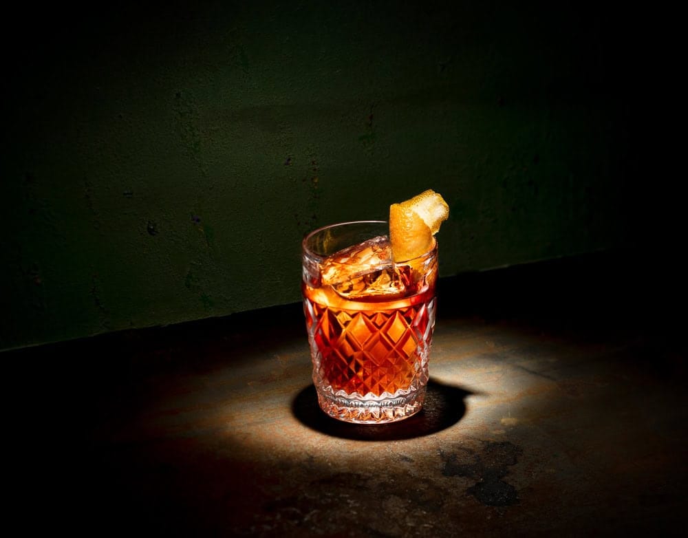 classic negroni dramatic lighting A classic Negroni cocktail with an orange twist, captured under dramatic lighting against a textured green backdrop.