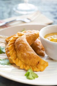 Three fried empanadas served with a bowl of creamy dipping sauce on a white plate, garnished with fresh cilantro.