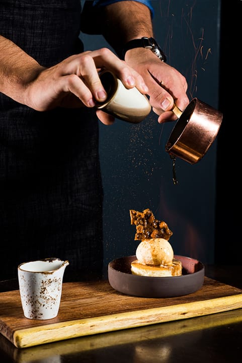 Flaming tres leches cake topped with ice cream and a caramelized tuile, being drizzled with caramel sauce.