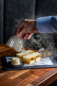 Focaccia with smoked butter and tapenade, served with a glass dome on a dark plate in a smoky presentation.