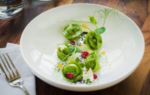 Green Tortellini Pasta Bowl – Food Photography Green tortellini pasta in a white bowl with garnishes on a wooden table.