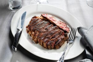 Grilled ribeye steak served on a white plate with a knife and fork, accompanied by a drink.