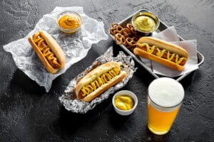 Hot Dogs with Pretzels and Beer | Food Photography Three hot dogs with mustard, served with pretzels, mustard dips, and a glass of beer on a dark surface.