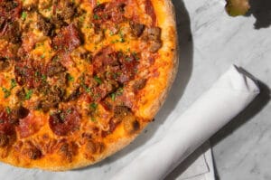 Meat lovers pizza topped with various meats and herbs on a marble surface & white napkin roll.