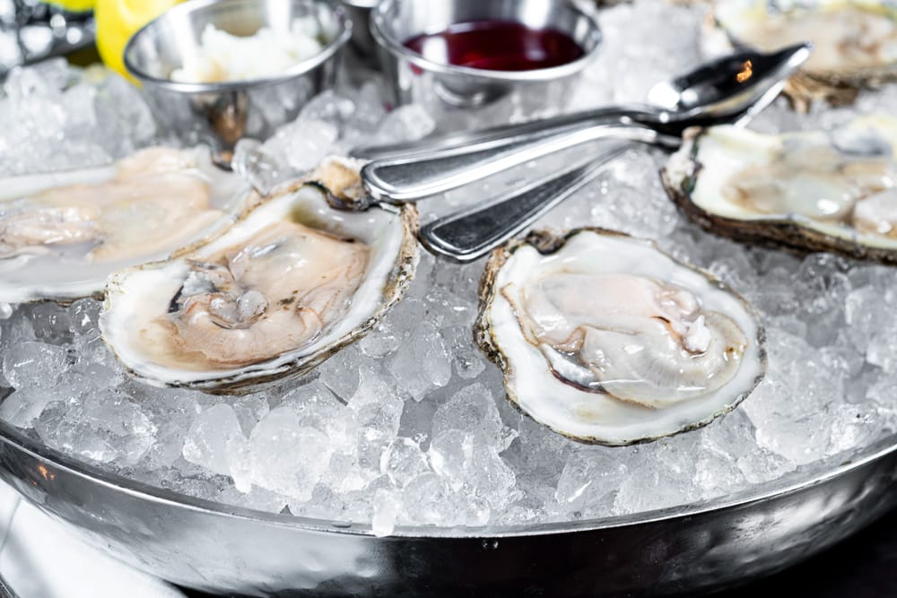 oysters-half-shell-ice Fresh oysters on the half shell served on ice with lemon wedges and dipping sauces in a metal dish.