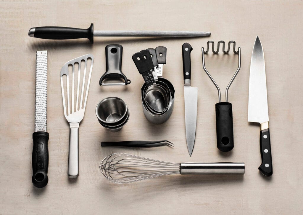 A flat-lay composition of professional chef tools, including knives, measuring cups, graters, and whisks.