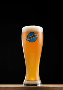 A chilled Blue Moon pint glass on a black background, showcasing vibrant amber beer with a foamy head.